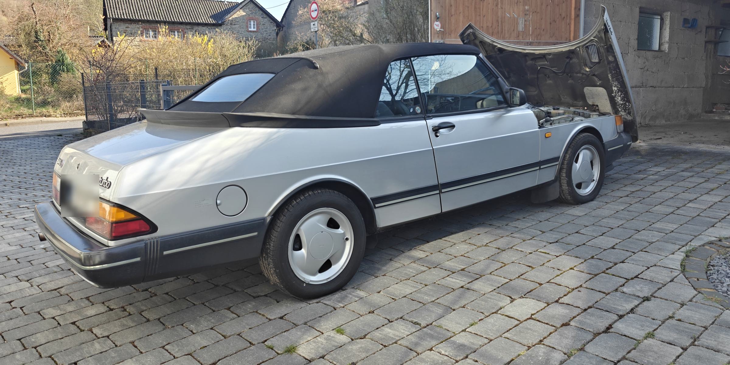 Silberner Saab 900 Turbo Cabriolet mit geöffneter Motorhaube auf einem gepflasterten Hof